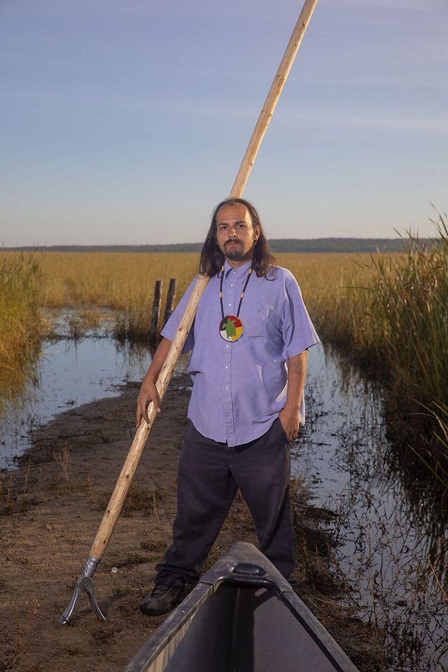 A man stands near a canoe with a long pole used to push it through the water.