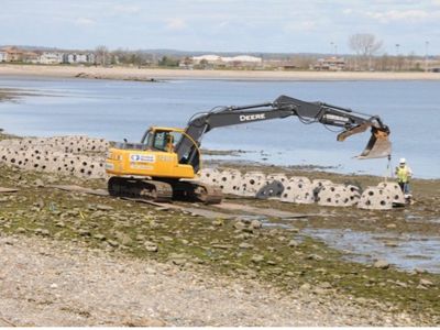 A yellow excavator places concrete reef balls along a shoreline.